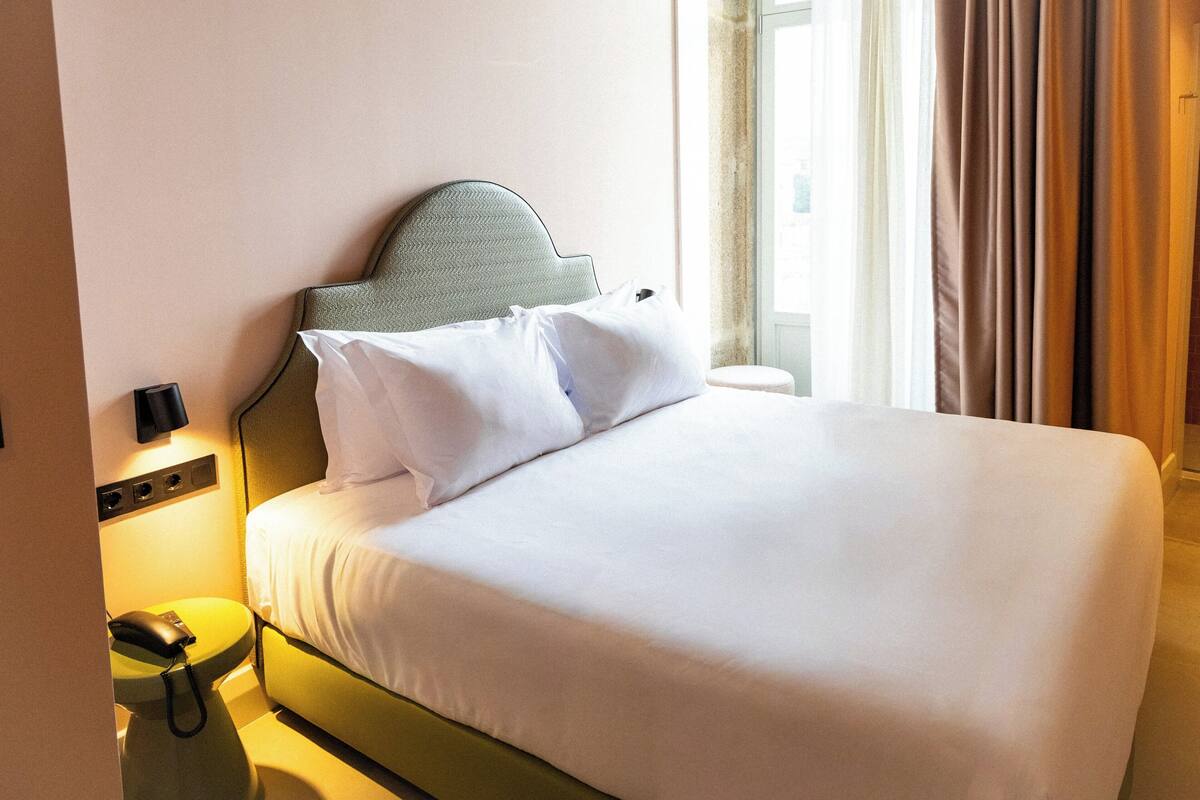 A hotel room with a bed, a bedside table with a phone, and a window with curtains.