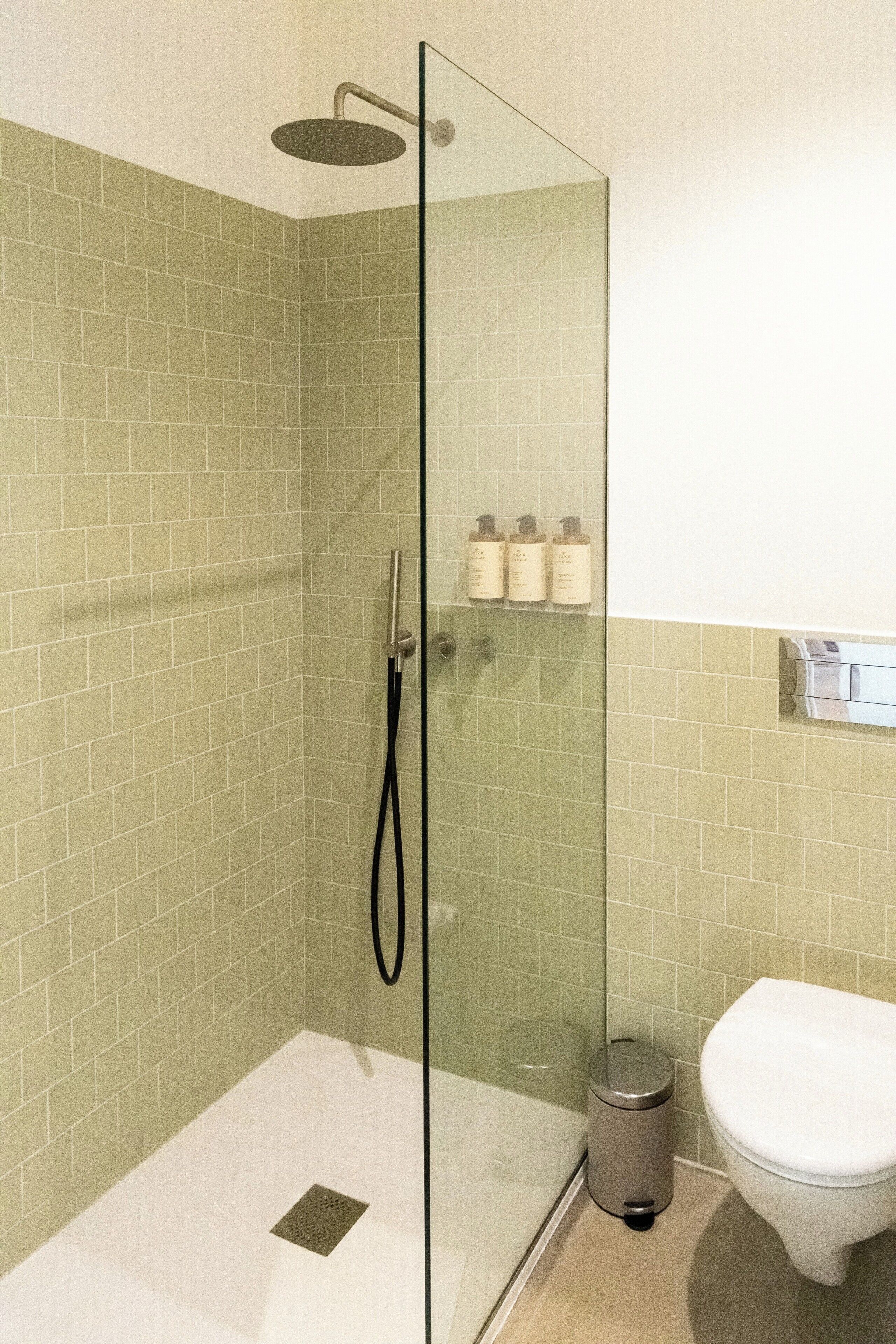 A glass-enclosed shower with a handheld showerhead, a toilet, and a trash can in a bathroom with green-tiled walls.
