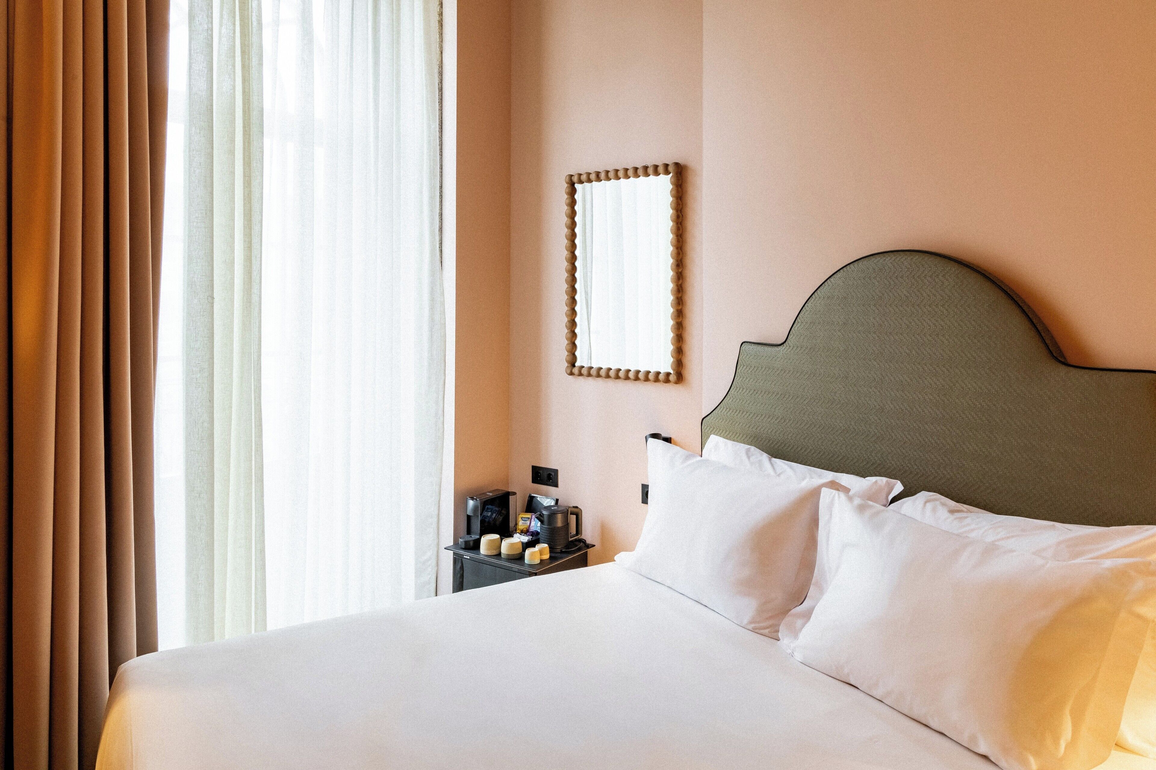 A hotel room with a bed, a nightstand with a lamp, a mirror, and a window with curtains.