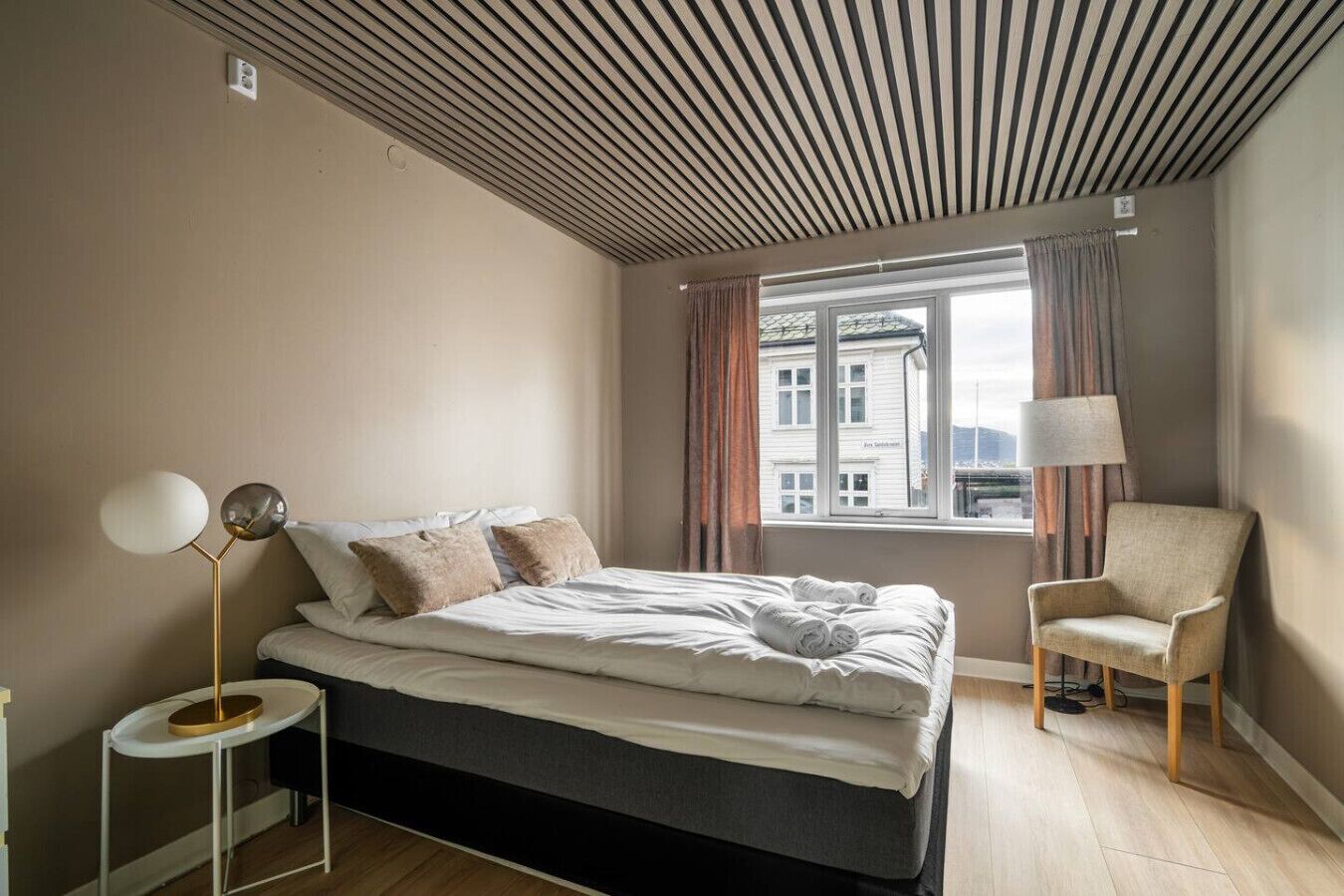 Un dormitorio moderno con cama, lámpara de noche, silla y vista a un edificio desde la ventana.