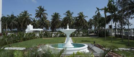 A white fountain surrounded by a well-maintained garden with palm trees and a clear blue sky.