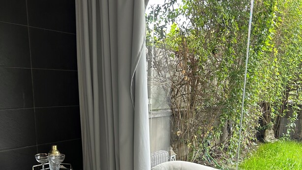 Um quarto com piso de mármore, um pilar branco com topo de vidro, uma cadeira cinza, uma cama com cabeceira branca e vista para o verde lá fora.