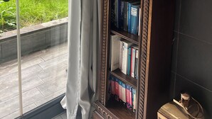 Um quarto com uma grande janela, uma estante com livros, um busto e um espelho fixado na parede.