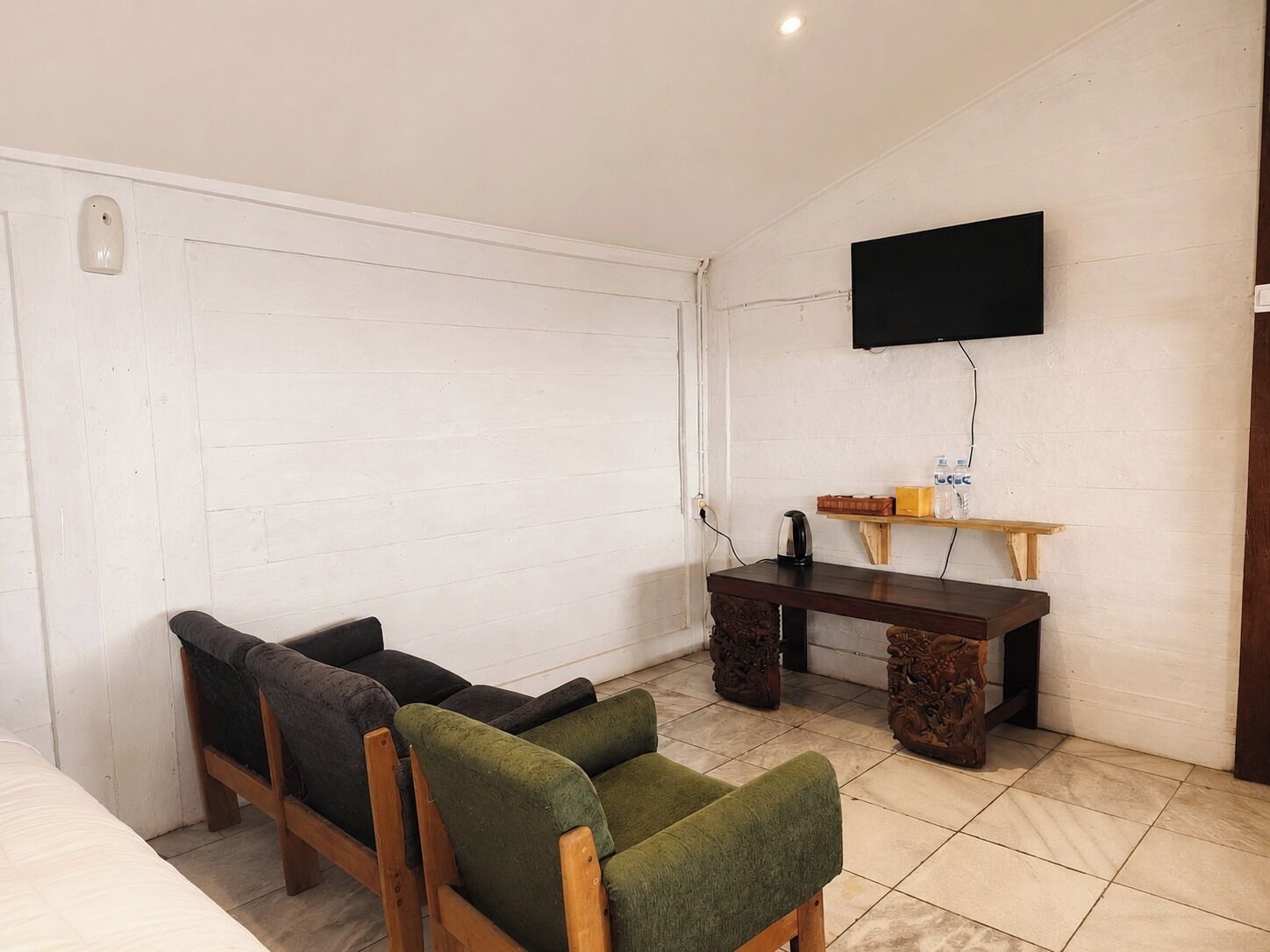 A room with a bed, a sofa, a wooden table, a television mounted on the wall, and a small shelf with items on it.