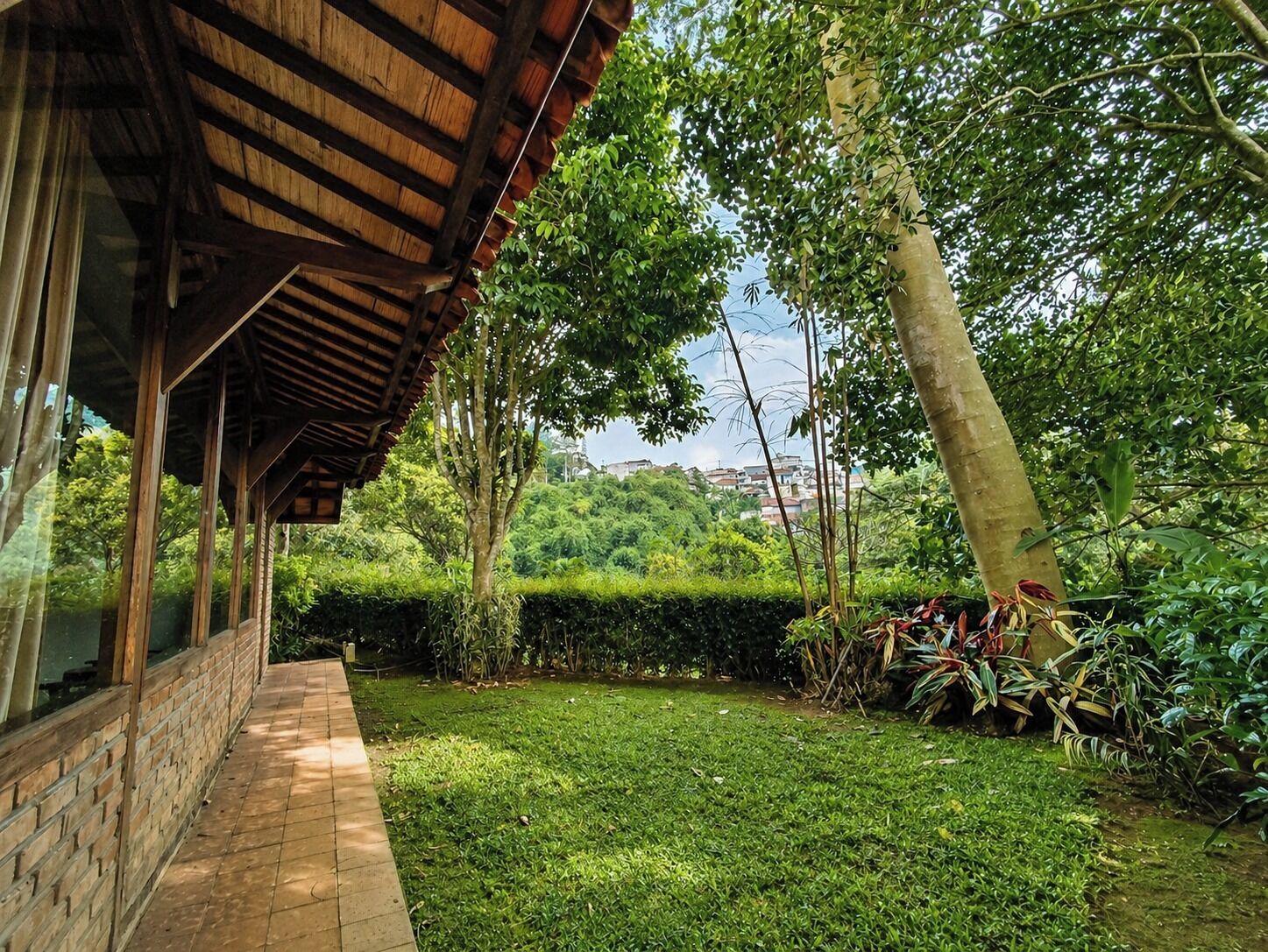 A covered outdoor area with a brick pathway leading to a lush green lawn, surrounded by trees and a view of a distant town.
