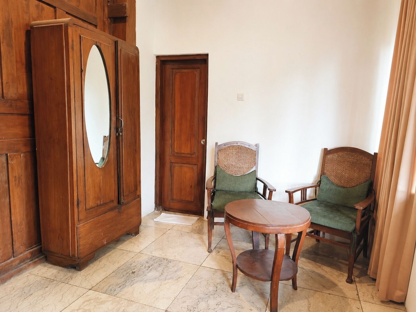 A room with a wooden wardrobe, a door, two green upholstered chairs, and a round wooden table.