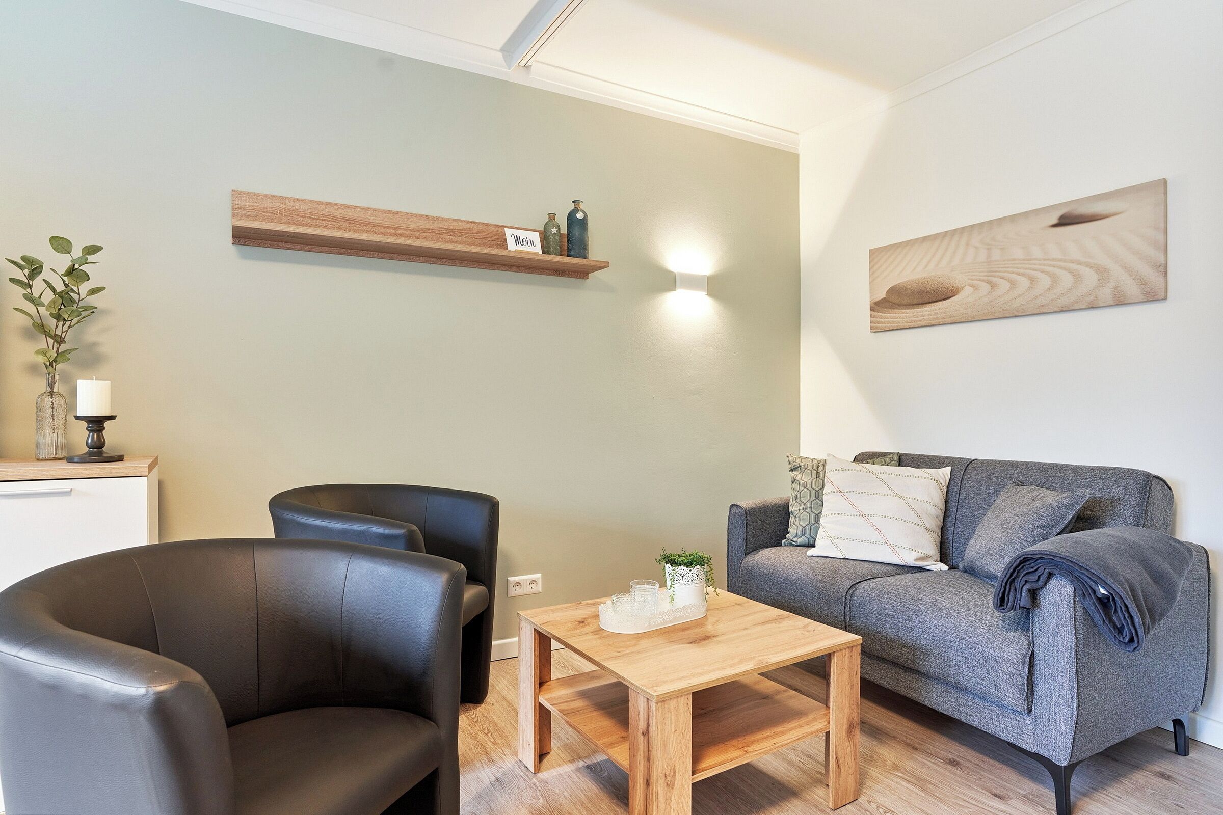 A modern living room with a grey sofa, a wooden coffee table, and a black leather armchair. A wooden shelf with decorative items hangs on the wall, and a painting is displayed above the sofa.