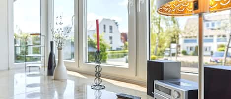 Una sala de estar moderna con un ventanal grande, un jarrón blanco con ramas, una vela alta, un equipo estéreo Philips y un control remoto sobre una encimera de mármol.