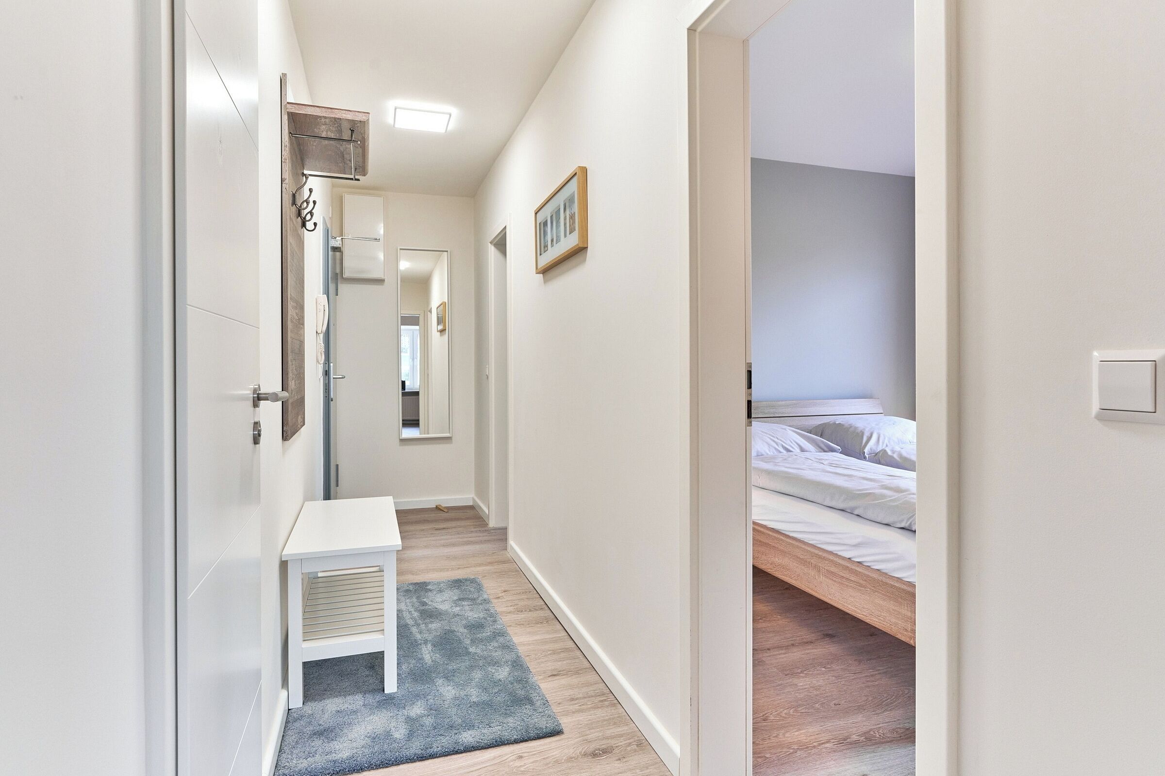 Un couloir étroit avec un bureau blanc et un tapis bleu, qui mène à une chambre à coucher avec un lit en bois et du linge de lit blanc.