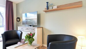 Una sala de estar moderna con un televisor de pantalla plana, dos sillas negras, una mesa de centro de madera y un florero con flores.