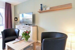 Ein modernes Wohnzimmer mit Flachbildfernseher, zwei schwarzen Sesseln, einem Holztisch und einer Vase mit Blumen.