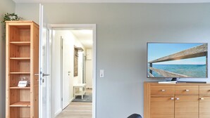 A room with a wooden cabinet, a television displaying a beach scene, and a doorway leading to another room. - House SeeZeit, Apartment 07 (Timmendorfer Strand)