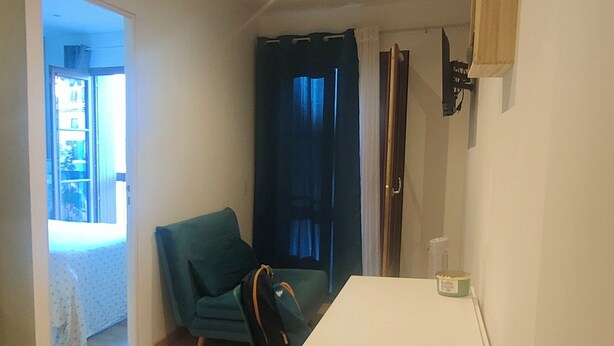 A small, well-lit room with a wooden floor, a white table, a green chair, and a black chair. There is a window with curtains, a door, and a shelf with items on the wall. - Le Derroja - Perpignan (Perpignan)