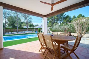 An outdoor patio with a wooden table and chairs, a ceiling fan, and a view of a swimming pool and garden. - Villa in lámetlla de Mar (Sant Jordi d'Alfama)