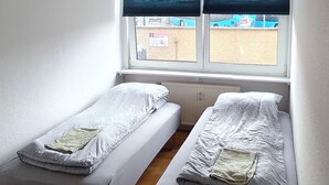 A small, single-room with two single beds, a window with blinds, and a wooden floor.