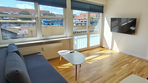 A modern living room with a sofa, a small table, and a TV mounted on the wall. Large windows offer a view of the street and buildings outside.