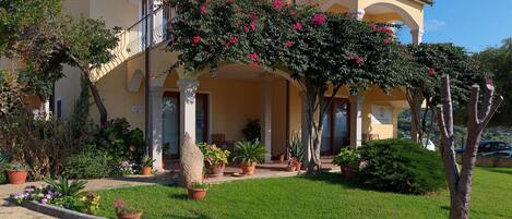 A two-story building with a yellow facade, a covered entrance with columns, and a balcony with pink flowering plants.