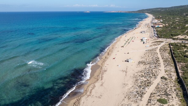 An aerial view of a coastal beach with clear blue waters, sandy shore, and a few buildings along the coastline. - Comfy Accommodation With sea View (Badesi)