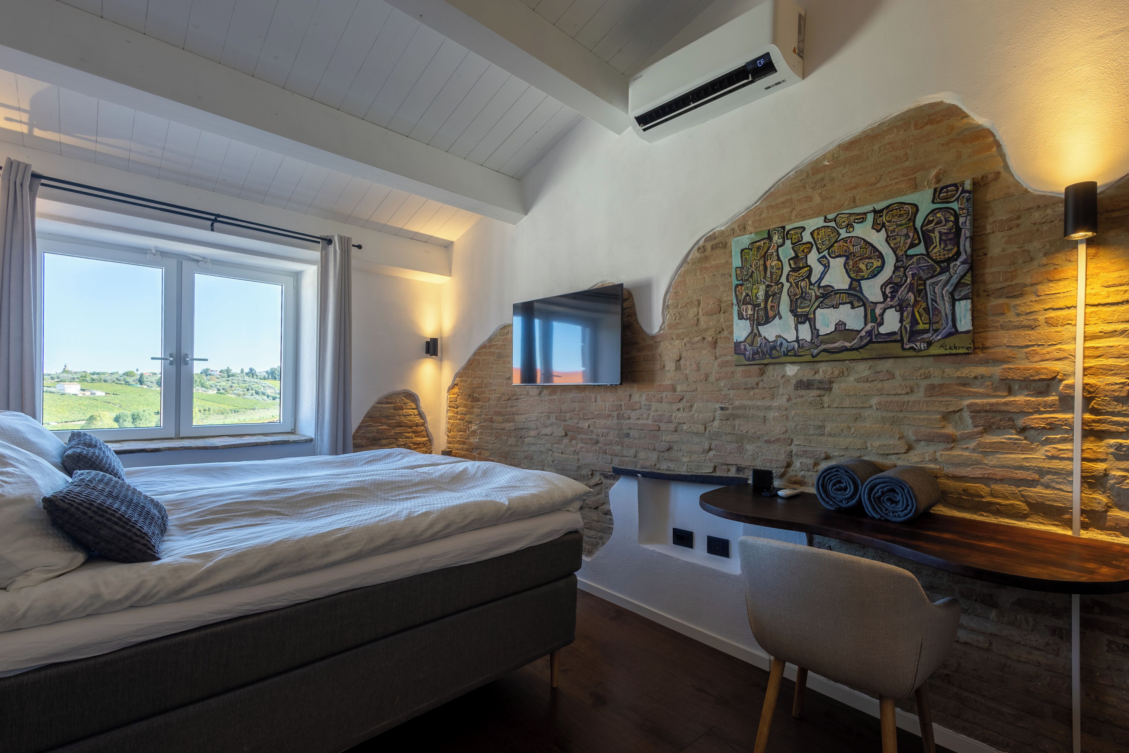 Un dormitorio con cama, televisor montado en una pared de piedra, escritorio con silla y vista al exterior.