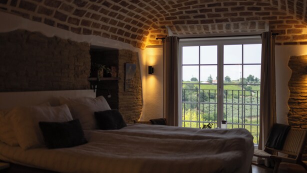 A bedroom with a large bed, a chair, a window with a view of the countryside, and a brick ceiling.