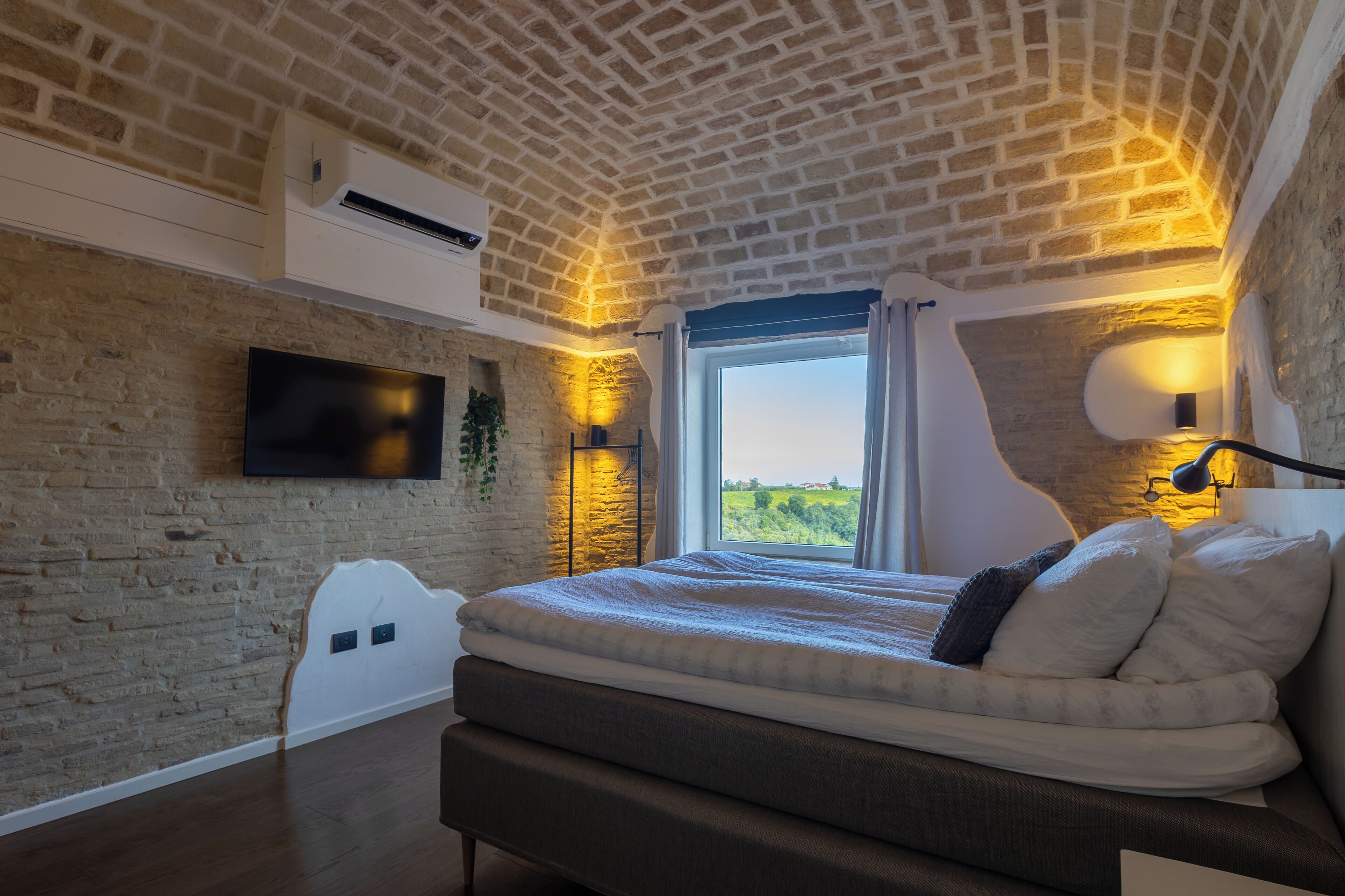 A bedroom with a brick ceiling, a flat-screen TV mounted on the wall, a large window with curtains, and a bedside lamp.