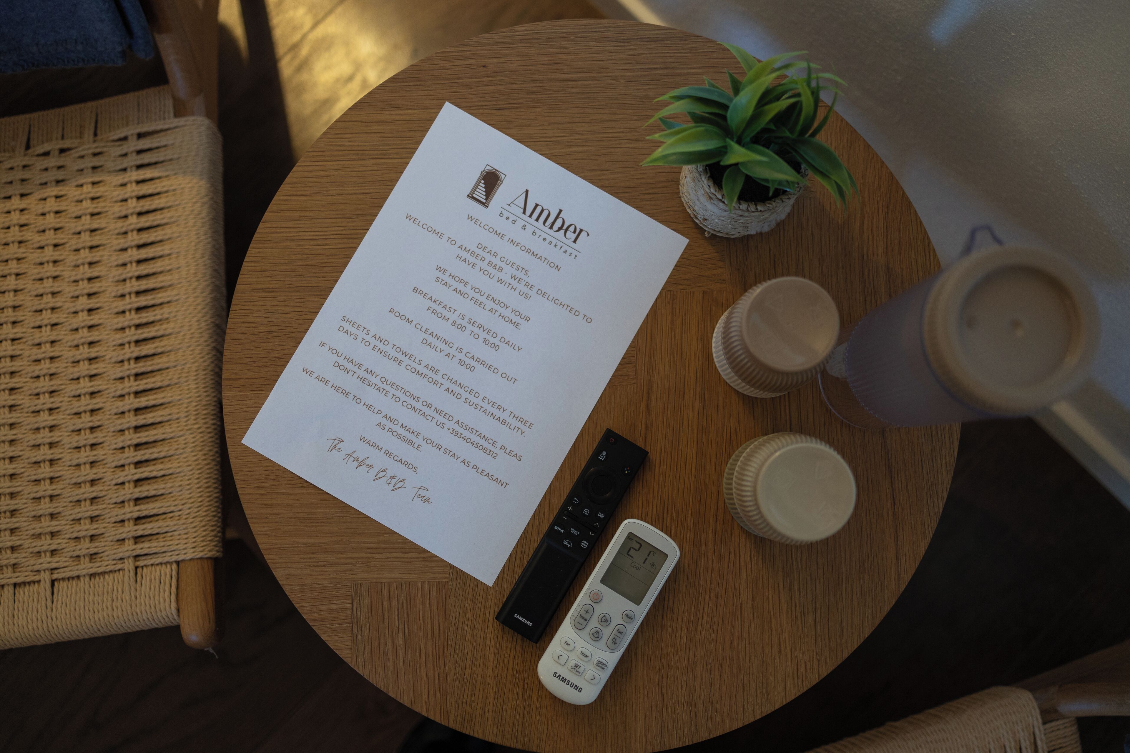Een ronde houten tafel met een afstandsbediening, een Samsung telefoon, een kleine potplant en een welkomstkaart voor hotel Amber.