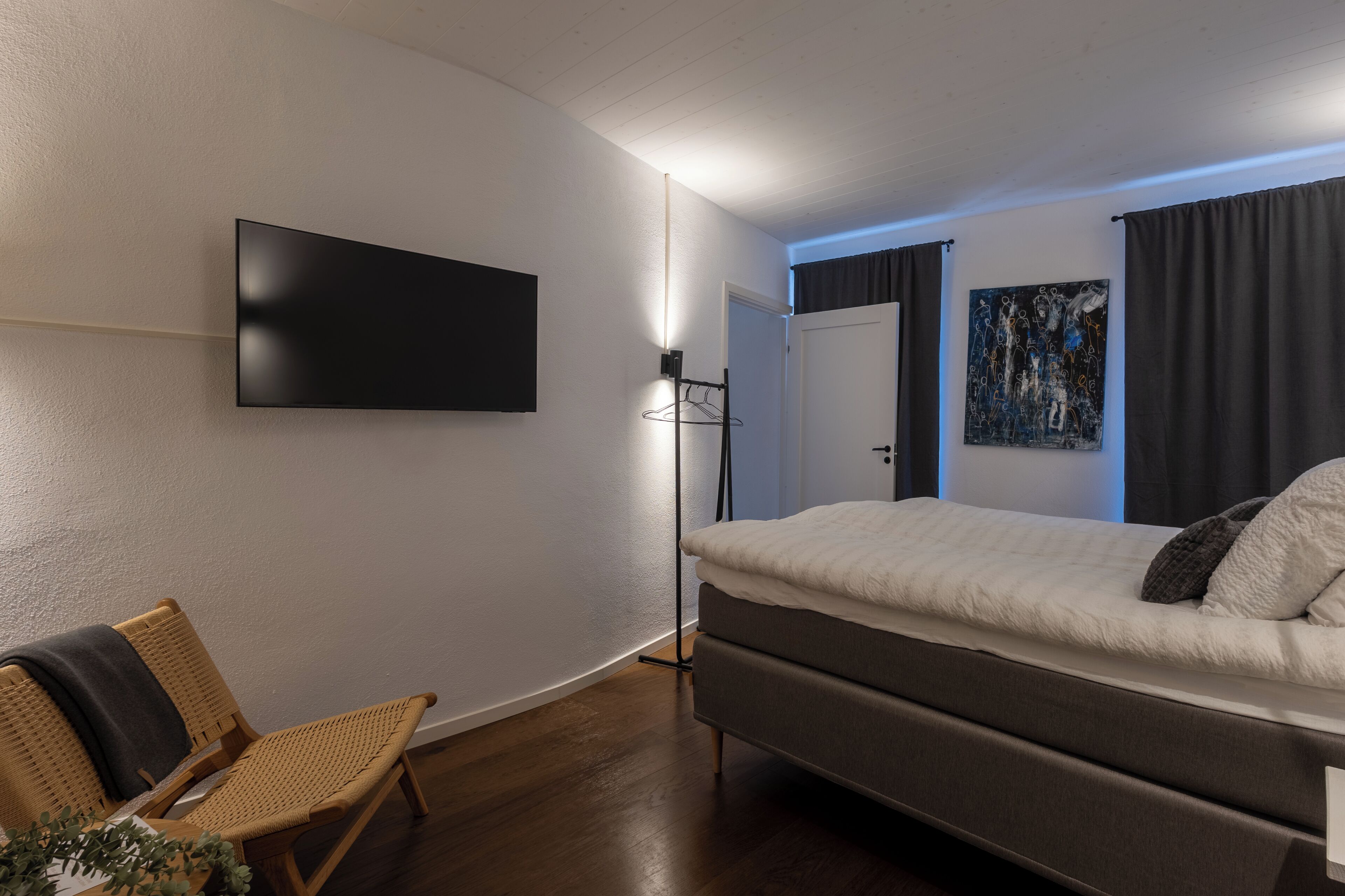 Une chambre à coucher moderne avec un lit, un téléviseur fixé au mur, une chaise et un tableau accroché au mur.