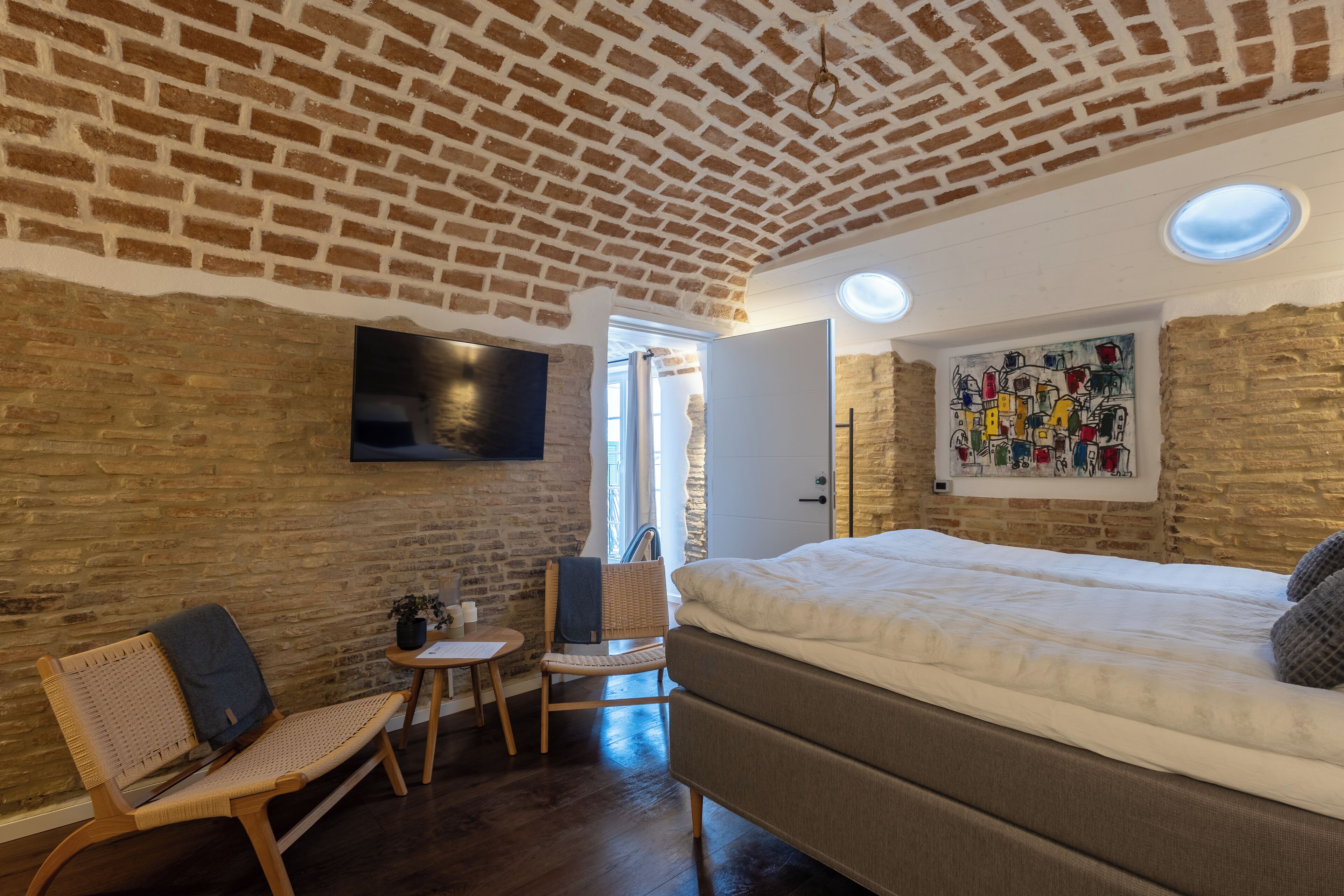 A room with a brick ceiling, a bed, a chair, a small table, and a painting on the wall.