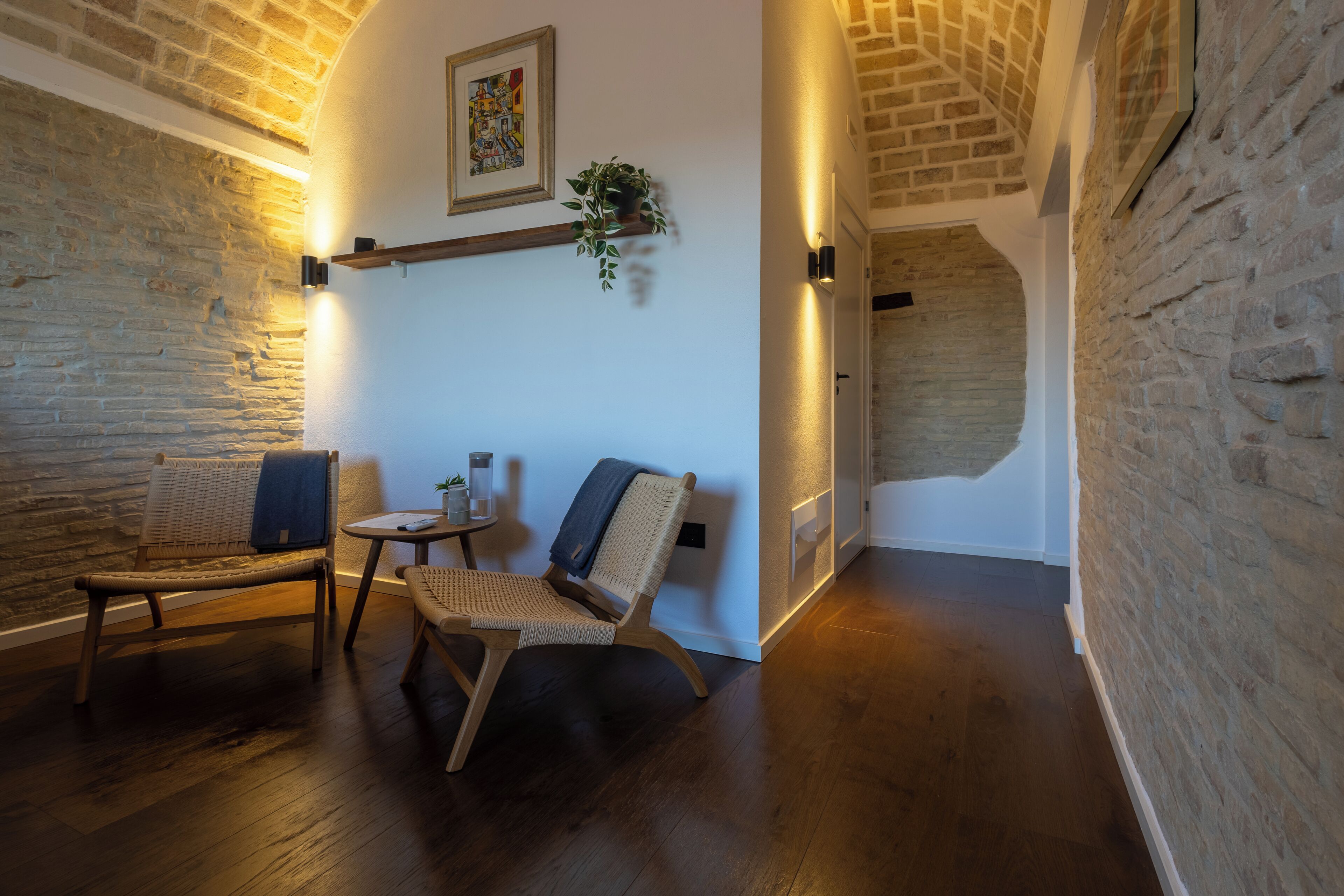 Ein gemütliches Zimmer mit Holzboden, zwei Korbstühle, einem kleinen Tisch, einer Pflanze und einem gerahmten Kunstwerk an der Wand.