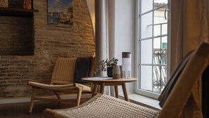 A cozy room with a brick wall, a wooden chair, a small round table with a vase and a candle, and a window with a view of the outside.