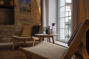 Una habitación acogedora con pared de ladrillo, una silla de madera, una pequeña mesa redonda con un florero y una vela, y una ventana con vistas al exterior.