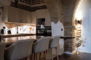 Una cocina con armarios blancos, encimera de madera y pared de piedra. Hay dos taburetes de bar y un florero con flores sobre la encimera.