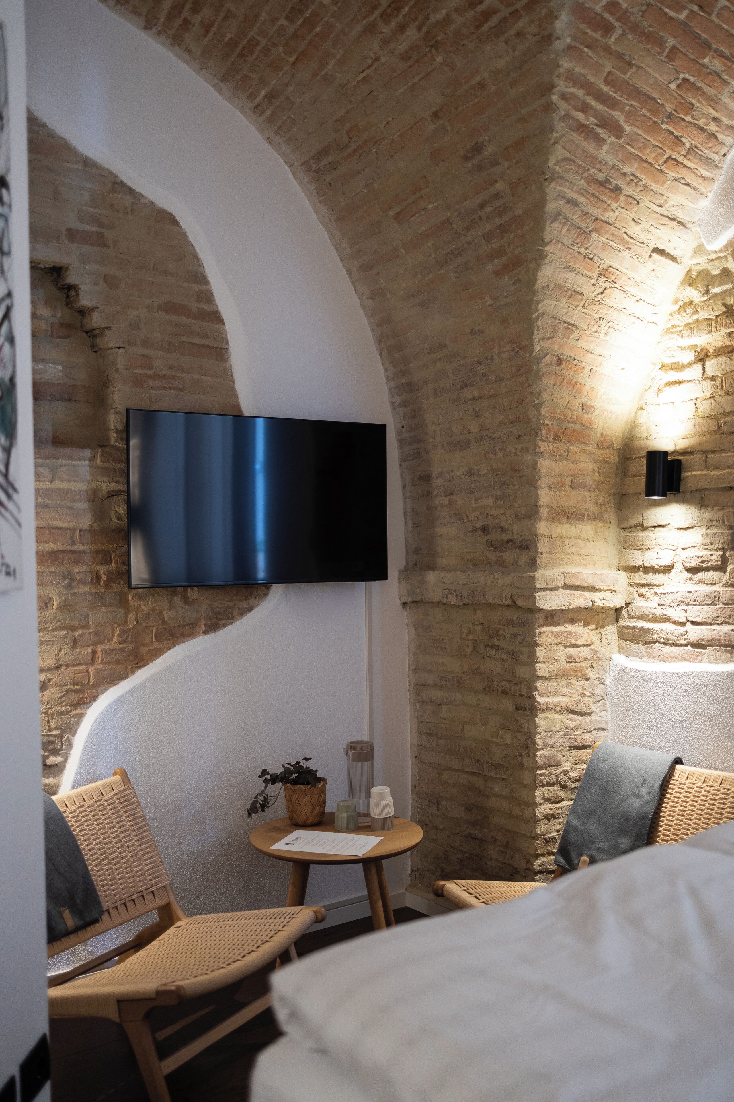 A cozy room with a brick wall, a flat-screen TV mounted on the wall, wicker chairs, a small round table with a candle and a plant, and a bed with a white headboard.