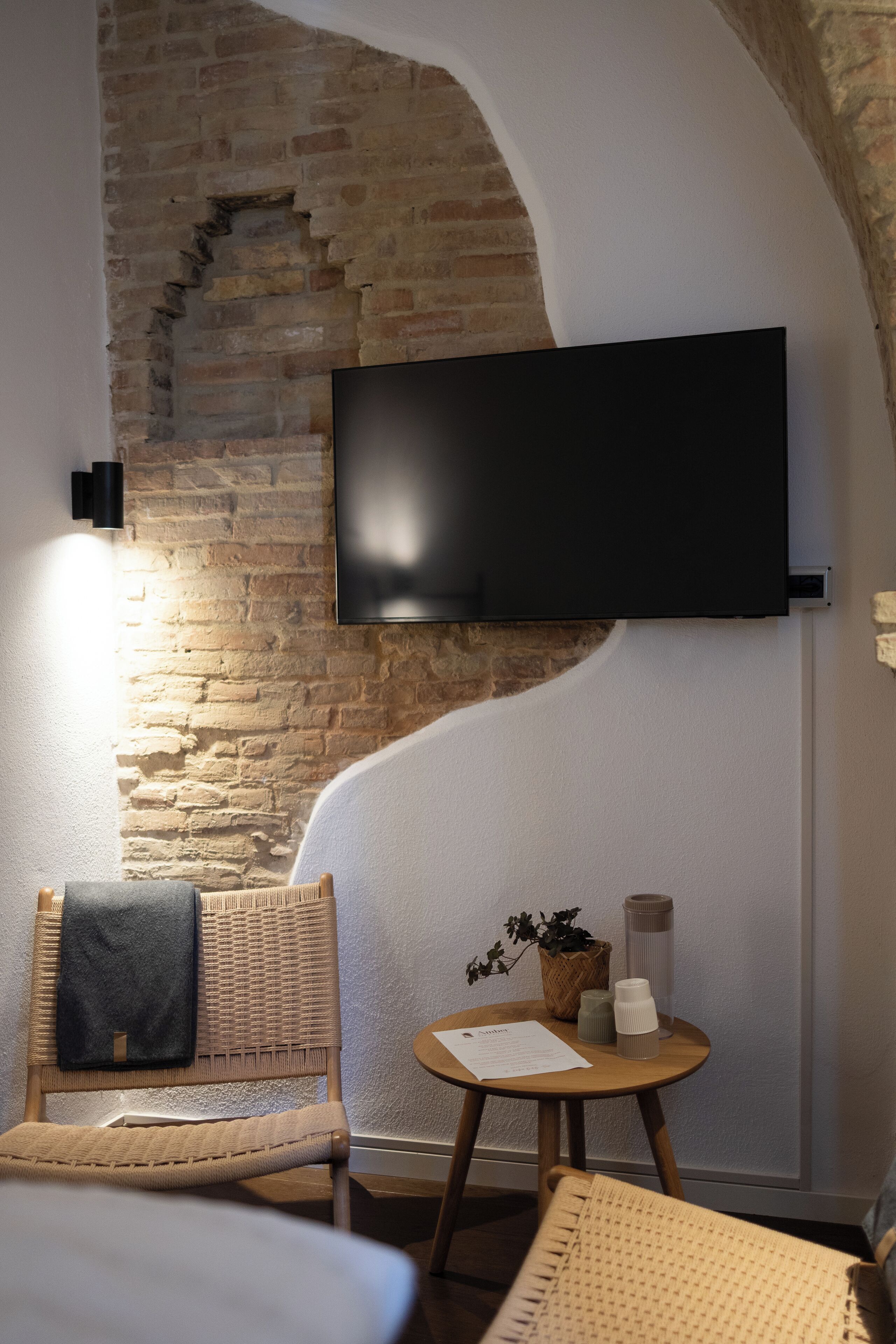 Una habitación acogedora con pared de ladrillo, un televisor de pantalla plana sobre una silla de mimbre, una pequeña mesa redonda con una planta y una toalla colgada del respaldo de la silla.