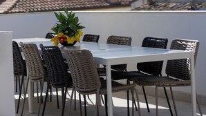 An outdoor patio with a white table and wicker chairs, an umbrella, and a view of buildings and hills in the background.