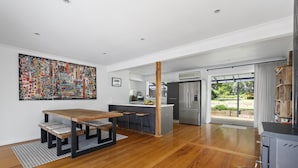 A modern kitchen with a large island, a dining area with a wooden table, and a view of the outdoors through a sliding door. - Cooinda, Red Hill South - Serene Red Hill South Retreat surrounded by gardens. (Red Hill South)