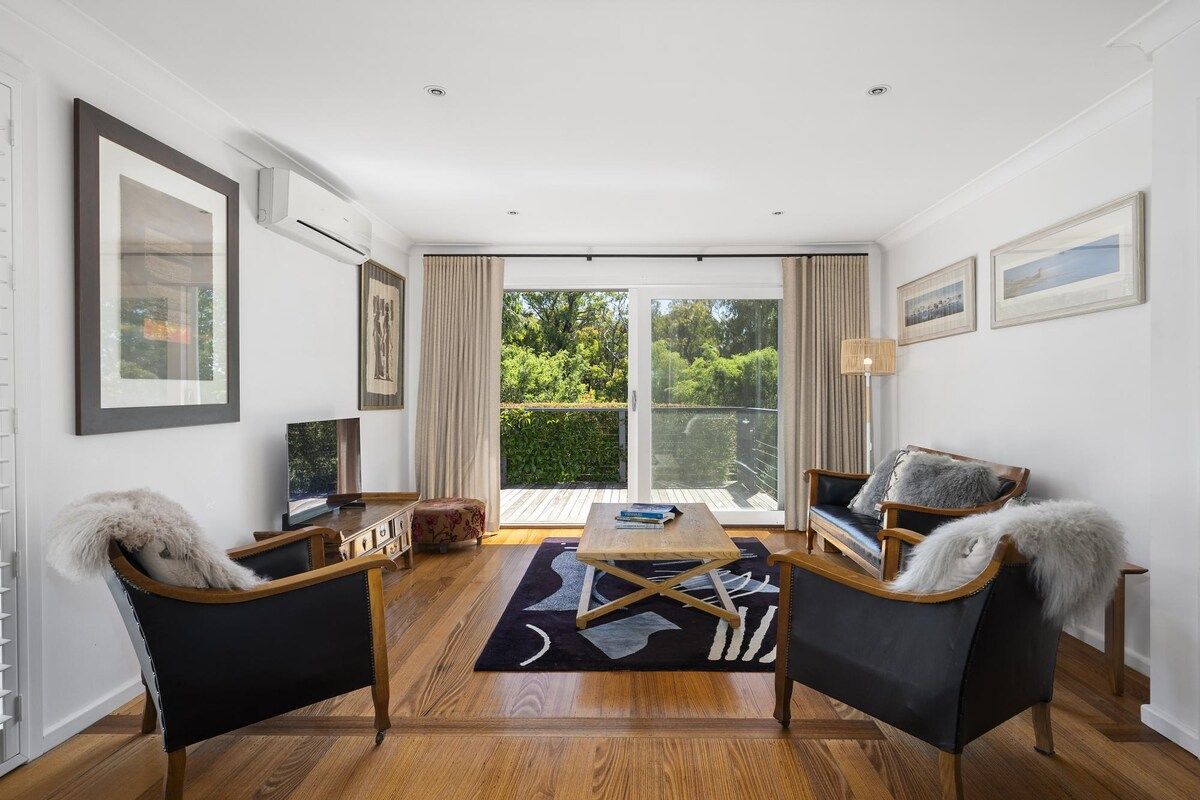 Un salon moderne avec un plancher en bois, deux canapés en cuir noir, une table basse et une grande fenêtre donnant sur un paysage verdoyant.