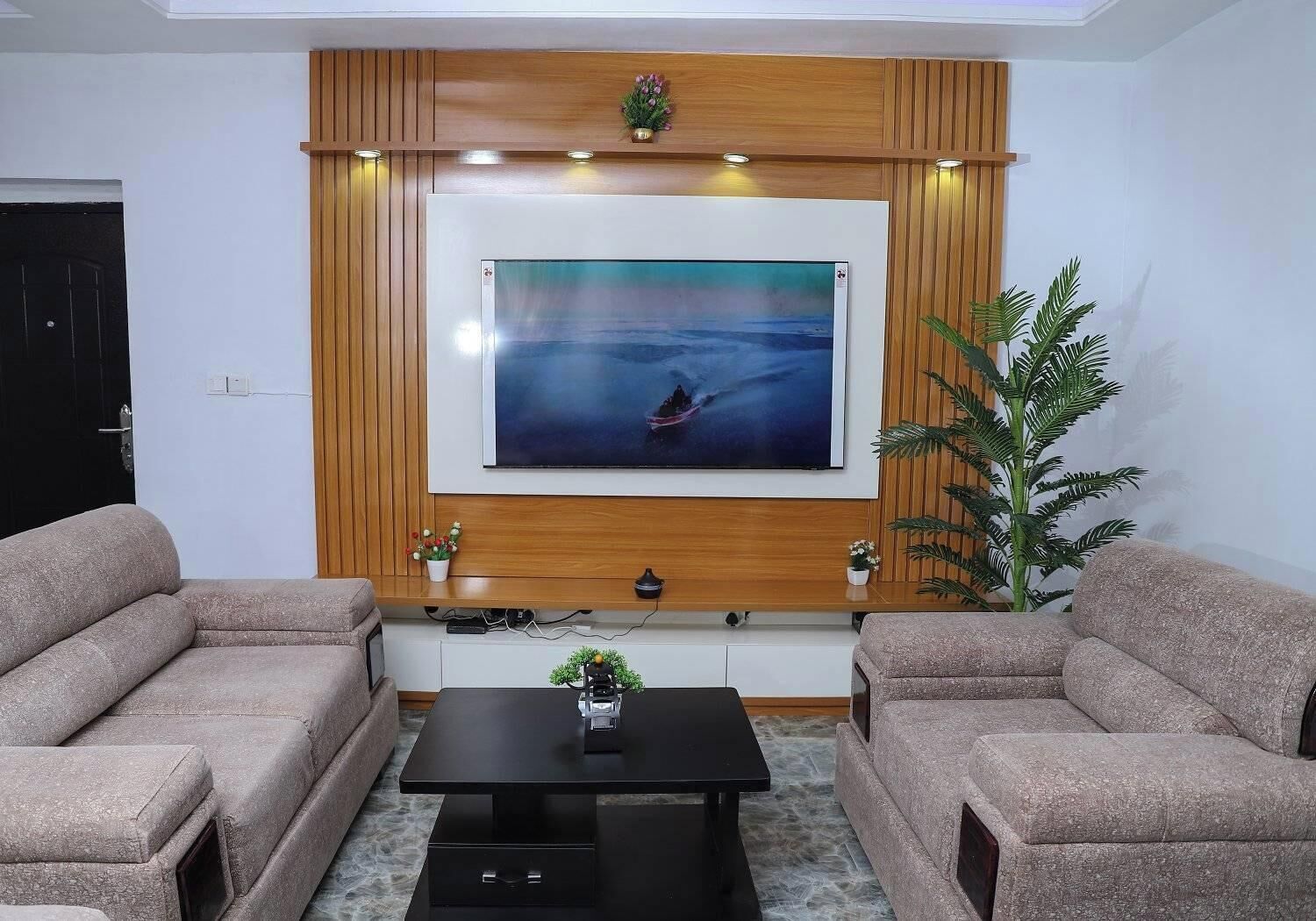 A living room with a large flat-screen TV mounted on a wooden wall, two beige sofas, a black coffee table, and a potted plant.