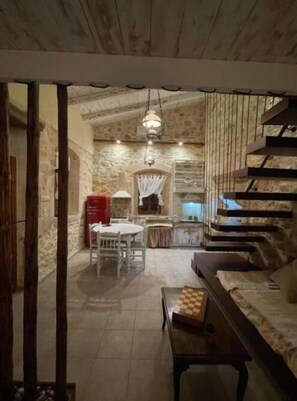 A rustic interior with a stone wall, a wooden staircase, a dining area with a table and chairs, and a hanging light fixture.