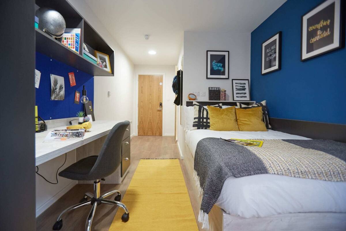 A bedroom with a bed, a desk, and a chair. There are wall decorations and a door in the background.