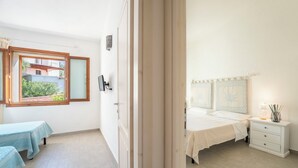 A bedroom with a bed, bedside table, and a window with a view of a building and greenery. - Lovely Flat Immersed in Nature (Badesi)