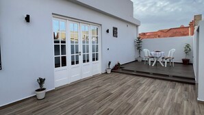 Un edificio moderno con techo plano, paredes exteriores blancas y terraza de madera. Hay plantas en macetas y mobiliario exterior.