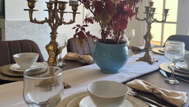 Una mesa de comedor dispuesta con platos, tazones y cubiertos blancos, un florero azul con hojas rojas y candelabros dorados.