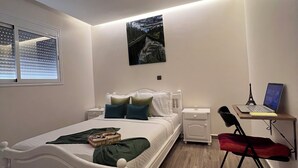 Un dormitorio con cama, escritorio con computadora portátil, silla, ventana con persianas y un cuadro en la pared.