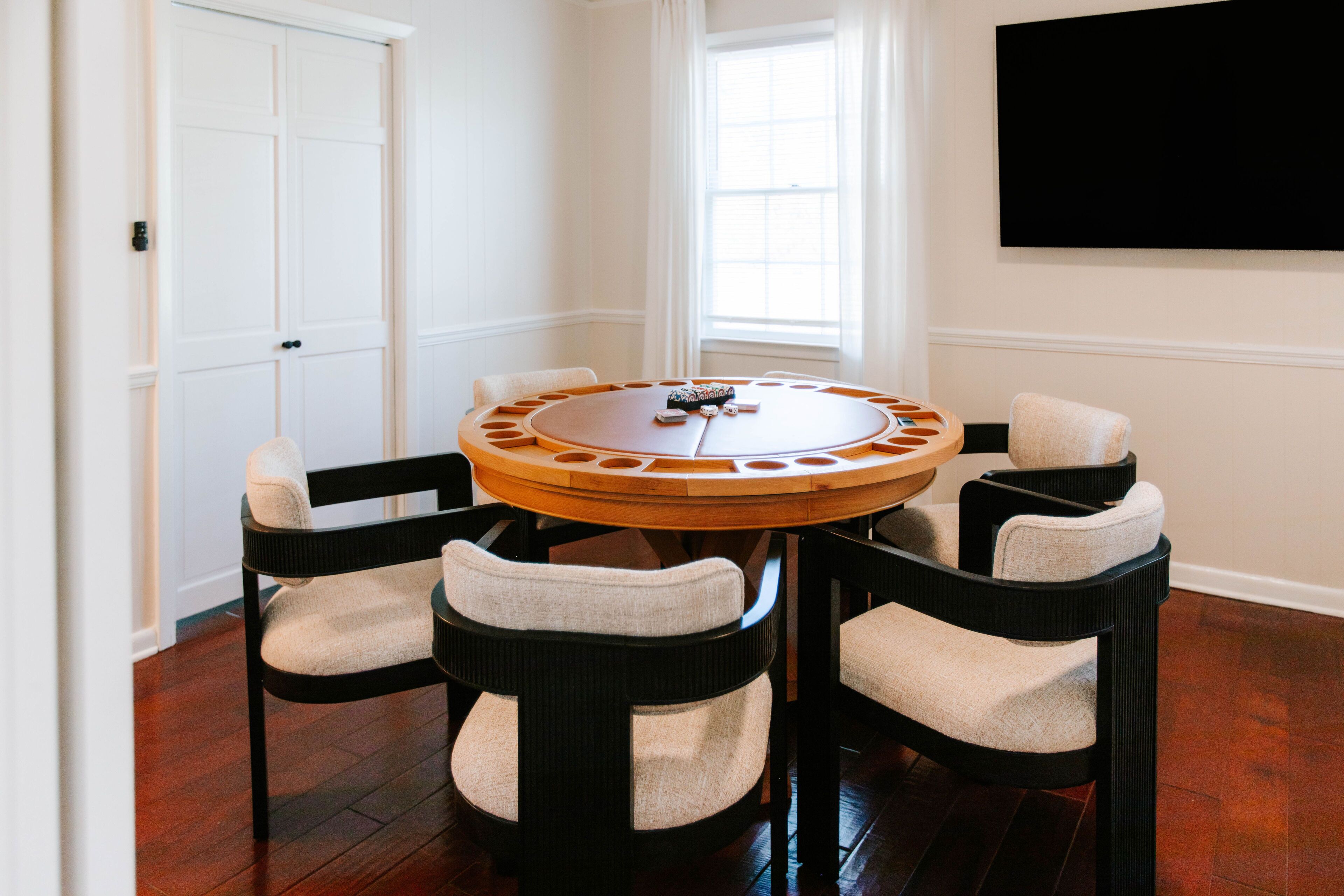 A round wooden table with chairs, a TV on the wall, and a door in the background.