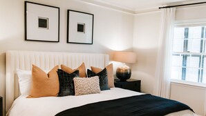 Ein ordentlich bezogenes Bett mit verschiedenen Kissen und einer schwarzen Tagesdecke. Über dem Bett hängen zwei gerahmte Bilder an der Wand.