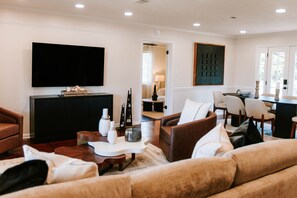 A modern living room with a flat-screen TV, comfortable seating, a dining area, and a hallway leading to another room.