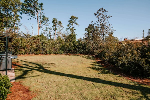 A grassy backyard with a barbecue grill, surrounded by dense shrubbery and tall trees.