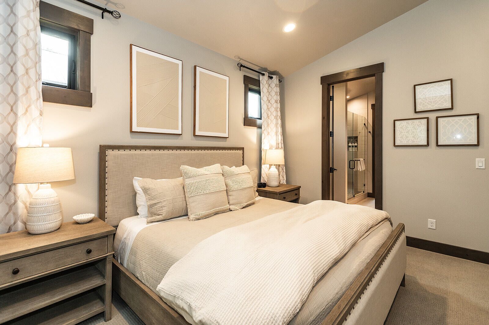 A bedroom with a large bed, two bedside tables with lamps, a window with curtains, and framed artwork on the wall.