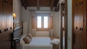A narrow room with a wooden ceiling, a bed with a white bedspread, a window with curtains, and wooden doors. - CASA RURAL & SPA LA GRAJA (Chinchón)